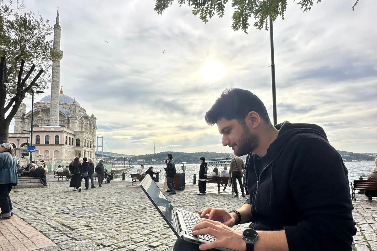 İlker Pehlivan, Ortaköy sahilinde bir bankta dizüstü bilgisayarla çalışırken; arka planda Ortaköy Camii ve Boğaz Köprüsü